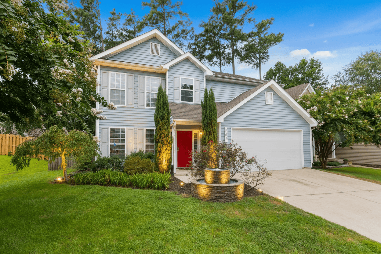 A light blue suburban house with a red front door, flanked by two tall, uplit evergreen trees and a glowing stone water feature.